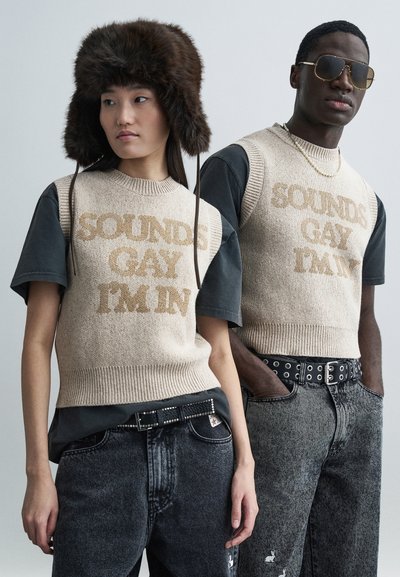 Beige gebreide trui met de tekst "SOUNDS GAY I'M IN", gecombineerd met zwarte t-shirts en vervaagde zwarte jeans. Een model draagt een bontmuts en een zonnebril.