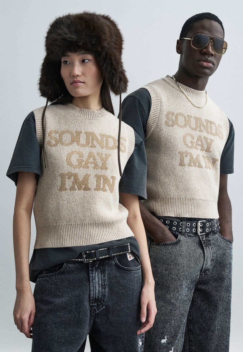 Μπεζ πλεκτό πουλόβερ με το κείμενο "SOUNDS GAY I'M IN", συνδυασμένο με μαύρα μπλουζάκια και ξεθωριασμένο μαύρο τζιν. Ένα μοντέλο φοράει γούνινο καπέλο και γυαλιά ηλίου.