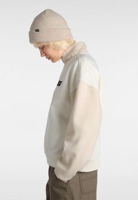 Bonnet en tricot côtelé beige et sweatshirt bicolore avec des couleurs beige clair et blanc cassé, avec un col zippé et un logo en patch sur la poitrine.