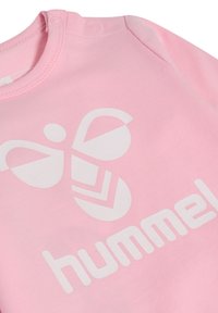 Lichtroze shirt met korte mouwen en witte logo met een gestileerd bijenontwerp, ronde halslijn en drukknoopsluiting op de schouder.