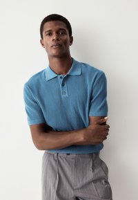 Turkos ribbad pikétröja med korta ärmar, utrustad med krage och tre knappar framtill, matchad med ljusgrå byxor med tunna ränder.