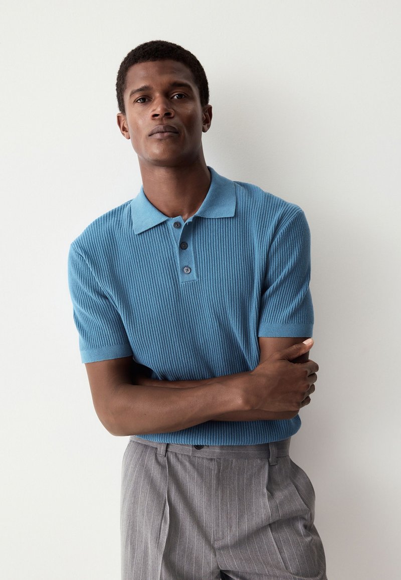 Turkos ribbad pikétröja med korta ärmar, utrustad med krage och tre knappar framtill, matchad med ljusgrå byxor med tunna ränder.