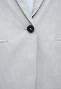 Veste de costume gris clair boutonnée avec un seul bouton foncé, montrant des revers et deux poches avant.