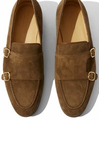 Bruine suede loafers met dubbele gouden gespen, een gladde textuur, ronde neus en contrasterende stiksels langs de randen. Klassiek instapmodel.