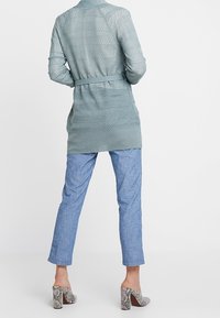 Ljusgrön stickad kofta med knytbälte, bars över ljusblå avsmalnande byxor och mönstrade skor med klossklack. Texturerat tyg med öppet stickmönster.