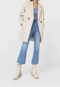 Manteau beige à double boutonnage, débardeur court bleu clair, jean évasé taille haute, et bottines cr ème à talons épais. Textures lisses.