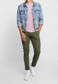 Jeansjacke, rot-weiß gestreiftes T-Shirt, olivgrüne Slim-Fit-Hose und weiße Turnschuhe mit gemusterten Socken. Legeres Outfit mit Lagen-Look.