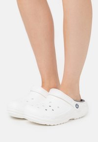 Zoccoli bianchi realizzati in materiale flessibile presentano fori di ventilazione, una suola strutturata e un rivestimento grigio a contrasto con un logo sul lato.
