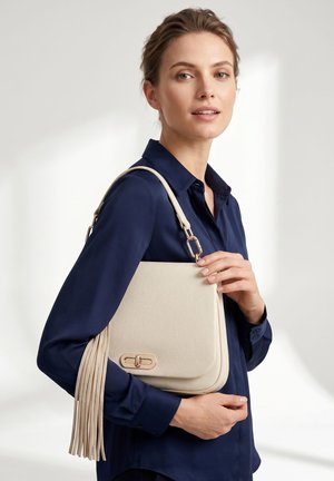 Femme en chemise bleu marine tenant un sac à main en cuir beige avec fermoir doré et bandoulière à pompon sur fond neutre.