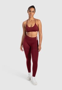 Burgundowe sportowe biustonosze i legginsy wysokiego stanu, wykonane z elastycznego materiału. Posiada cienkie ramiączka i logo na pasku. Białe sneakersy.