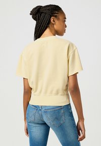 Felpa gialla a maniche corte con vestibilità comoda e orlo a costine, abbinata a jeans blu. Texture liscia, dettagli minimi sul tessuto.