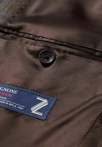 Tessuto marrone con una trama liscia presenta un bottone nero e un'etichetta blu con cuciture bianche, che indicano informazioni sul marchio e sulla cura.