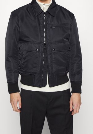 Schwarze Bomberjacke mit Reißverschluss, gerippten Bündchen und Saum sowie Klapptaschen mit Knöpfen vorne, getragen über weißem Pullover und schwarzen Hosen.