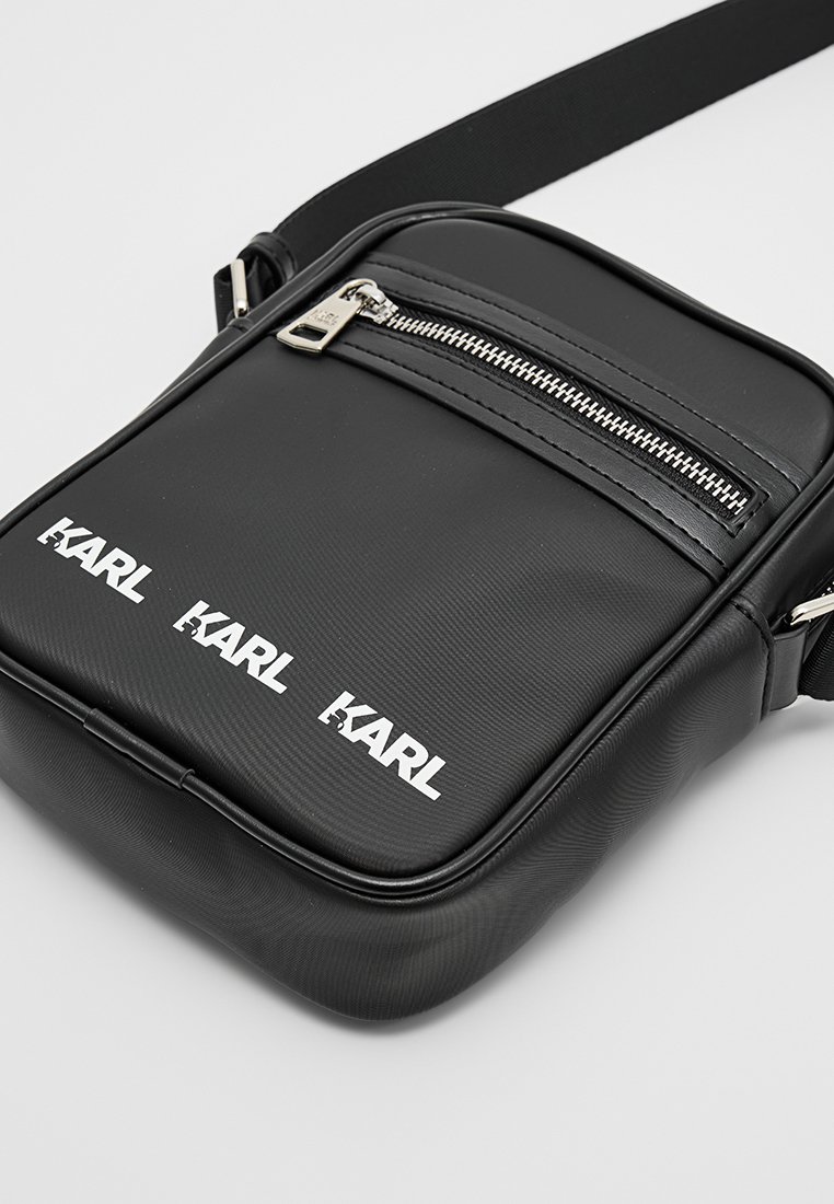 Bolso bandolera negro de cuero sintético liso, con un bolsillo delantero con cremallera, tres impresiones de logotipo blancas y una correa ajustable.