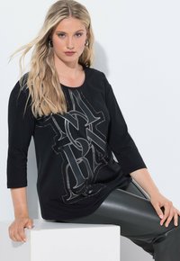 Schwarzes 3/4-Ärmel-Shirt mit einem großen dekorrativen silbernen Logo und Nieten-Details auf der Vorderseite. Weicher Stoff, lockere Passform.