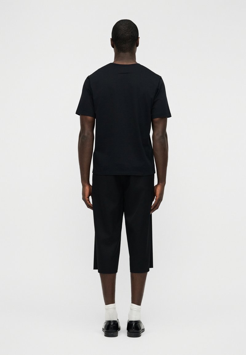 T-shirt en coton noir à manches courtes, associé à un pantalon court noir. La tenue présente un design minimaliste et une coupe décontractée. Des chaussures noires et des chaussettes blanches complètent le look.