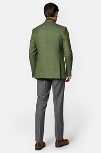 Zielona marynarka z wełny z domieszką, o dopasowanym kroju, z gładką fakturą, klapami i zapięciem na guziki przy rękawach. Połączona z szarymi spodniami.