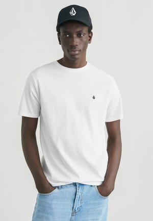 Camiseta blanca de algodón con cuello redondo y mangas cortas, combinada con vaqueros de mezclilla azul claro. Una gorra negra con un diseño de logo blanco.