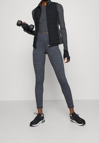 Svart väst med puffar över en grå långärmad topp, tillsammans med grå leggings. Håller mörka hantlar och har på sig svarta träningsskor.