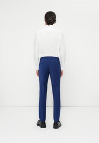 Pantalones de traje azul con una textura suave, ajuste slim y bolsillos traseros, combinados con una camisa blanca de botones, vistos desde atrás.