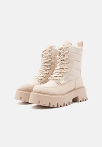 Χοντρές μπεζ μπότες μέχρι τον αστράγαλο με κορδόνια, με παχιές ραβδωτές σόλες και επενδεδυμένες πλισέ πλευρές σε άσπρο φόντο.