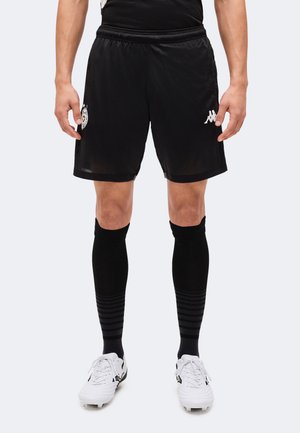 Modèle portant un short de football noir et des chaussettes montantes jusqu’aux genoux avec des chaussures de football blanches, sur un fond uni.