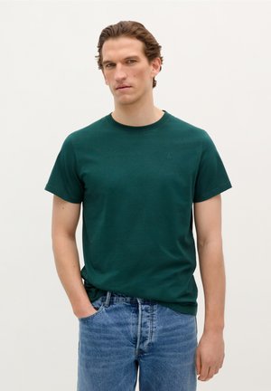 T-shirt en coton noir vert avec un col rond et des manches courtes. Doté d'un petit logo brodé sur la poitrine. Porté avec un jean en denim bleu.