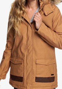 Chaqueta marrón con capucha, cierre frontal, dos grandes bolsillos y detalles en cuero marrón. La tela parece duradera con una textura suave.