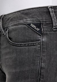 Jeans de denim gris oscuro con una etiqueta triangular negra con "REPLAY" en blanco, diseño de cinco bolsillos y una textura de desvanecimiento sutil.