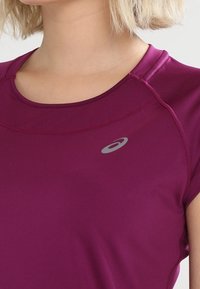 Kortärmad atletisk topp i djup magenta med rund halsringning och subtil textur; har en liten grå logotyp på framsidan.