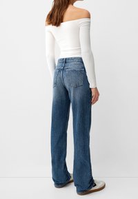 Mujer con blusa blanca de mangas largas y hombros al descubierto, jeans azules de tiro alto y sueltos, y zapatillas blancas, de pie frente a un fondo liso.