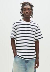 Jong persoon met dreadlocks, gekleed in een wit en zwart gestreept T-shirt met de tekst "Niet Storen" en lichte spijkerbroek, tegen een witte achtergrond.