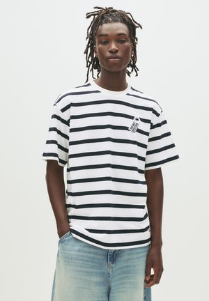 Ung person med dreadlocks iført en hvid og sort stribet t-shirt med teksten "Forstyrres ikke" og lyseblå jeans foran en hvid baggrund.