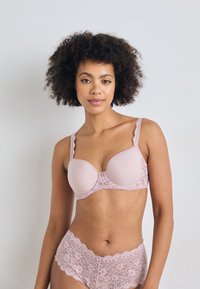 Woman with curly hair wearing matching light pink lace bra and briefs, standing against a plain white wall, facing forward.