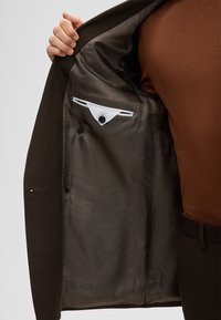 Binnenkant van een bruine colbert met een patroonvoering, een borstzak aan de linkerkant met een klep en twee knopen aan de voorkant.