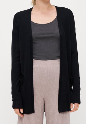 Femme portant un cardigan noir ouvert, un haut gris foncé à encolure dégagée et un pantalon côtelé beige taille haute, cadré des épaules aux hanches.