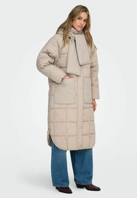 Beige vadderad rock med halsduk, med rektangulärt quiltat mönster, sidofickor och en midjelång modell. Kombinerad med jeans med vida ben.