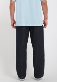Jean en denim bleu foncé avec une coupe décontractée, associé à un T-shirt bleu clair. Le look est complété par des baskets blanches avec des accents noirs.