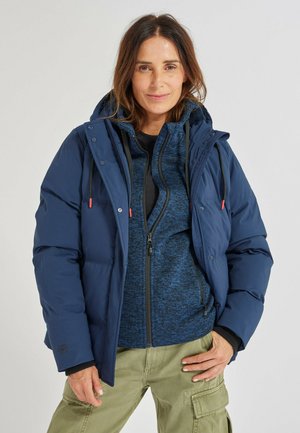 Mujer con forro polar azul de cremallera en capas y chaqueta acolchada con capucha azul marino, pantalones cargo oliva, posando frente a un fondo claro y sencillo.