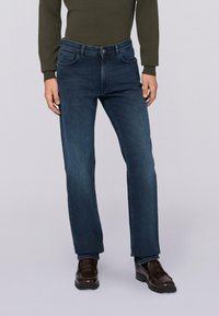 Hombre vistiendo unos jeans rectos de color azul oscuro, una camisa de manga larga de color verde oliva y unos mocasines de cuero marrón oscuro, de pie sobre un fondo claro.