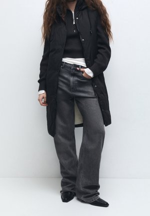 Kobieta ubrana w czarną długą pikowaną kamizelkę, czarny prążkowany sweter na białej bluzce, szerokie szare dżinsy oraz czarne spiczaste buty, stojąca na tle gładkiej ściany.