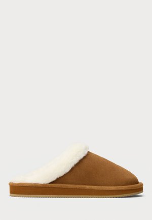 Bruna mockasin tofflor i mocka med vit fluffig foder och platt sula, sidovy från en enkel ljus bakgrund.