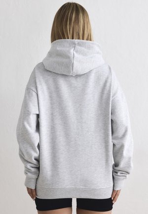 Person mit schulterlangem blondem Haar, die einen schlichten hellgrauen Kapuzenpullover und schwarze Shorts trägt, von hinten vor einem hellen Hintergrund gesehen.