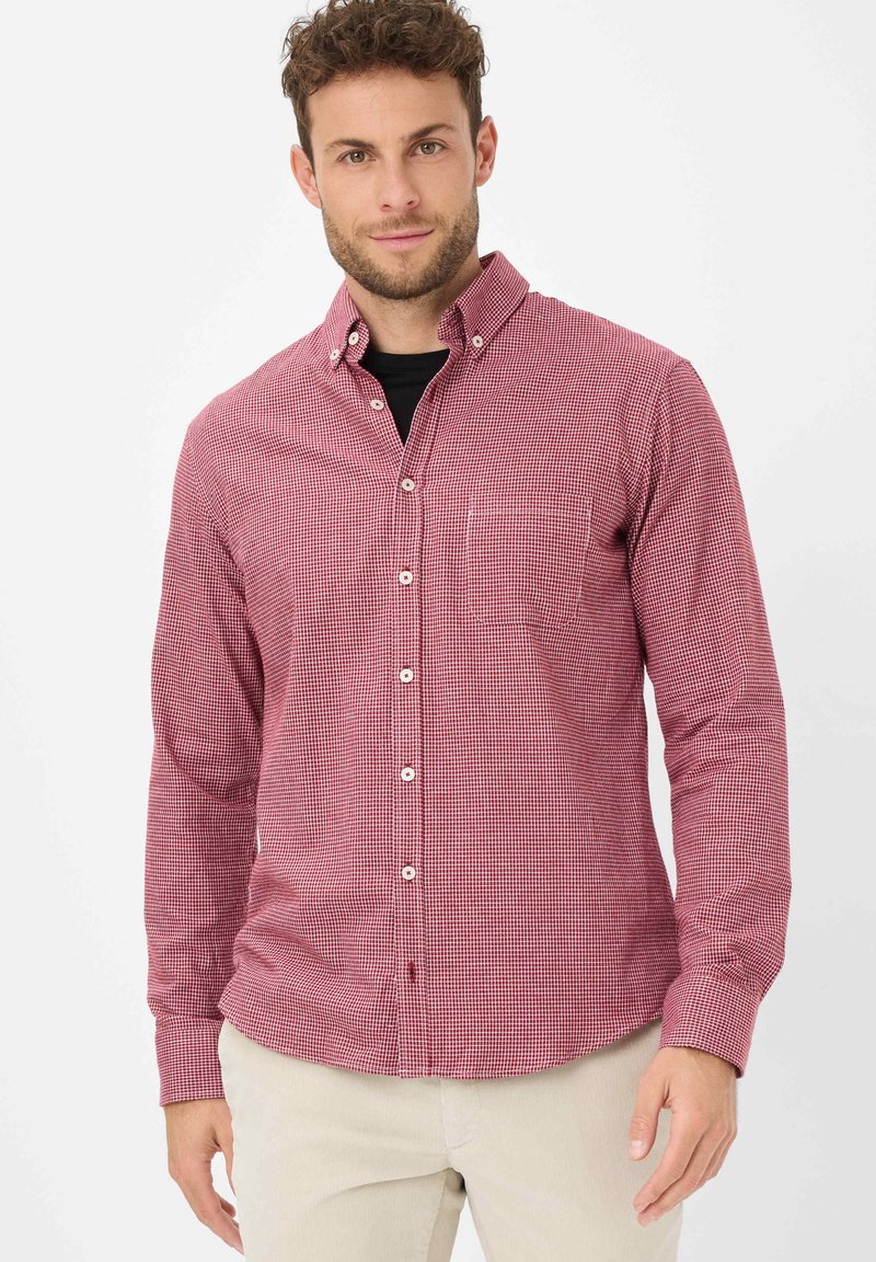 Uomo che indossa una camicia a quadri rossi e bianchi sopra una maglietta nera e pantaloni beige, in piedi su uno sfondo bianco semplice.