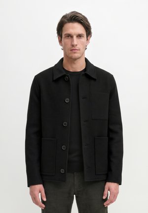 Homme portant une veste noire boutonnée avec trois poches, debout devant un fond blanc uni, regardant vers l'avant.