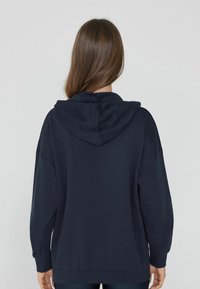 Navyblauer Hoodie mit einem geräumigen, lockeren Schnitt, einer zentralen Rückennaht, einer großen Kapuze und gerippten Bündchen und Saum. Weicher Textur.