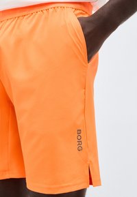 Φωτεινά πορτοκαλί σορτς με πλαϊνές τσέπες και λογότυπο "BORG" κοντά στο τελείωμα, φορεμένα από ένα άτομο με σκοτεινό δέρμα.