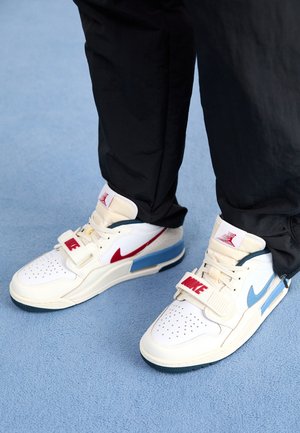 Osoba nosząca białe buty sportowe Nike z czerwonymi i niebieskimi akcentami, stojąca na jasnoniebieskim dywanie w czarnych spodniach.