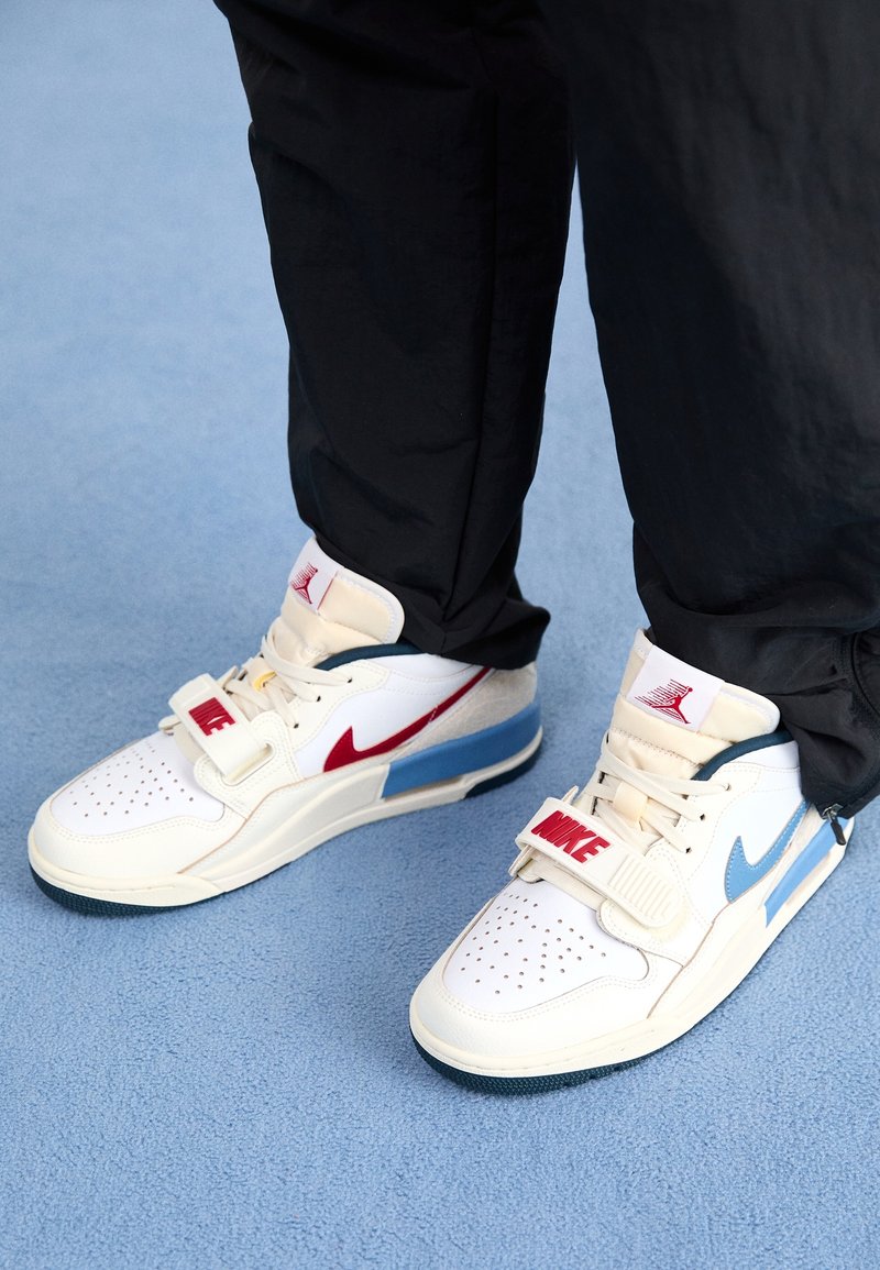 Personne portant des baskets blanches Nike avec des accents rouges et bleus, debout sur un tapis bleu clair avec un pantalon noir.