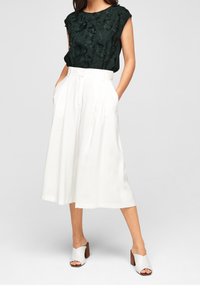 Tmavě zelený top s květinovými výšivkami, kombinovaný s bílými širokými kalhotami culottes. Doplněno bílými sandály na podpatku, které zdůrazňují čistou estetiku.